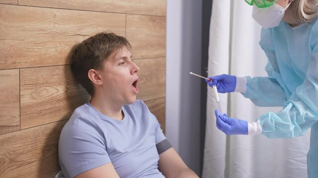 Health Professional In PPE Suit Introducing A Nasal And Throat Swab To Young Man Patient At Home Lying On Bed. Rapid Antigen Test Kit To Analyze Nasal Culture Sampling While Coronavirus Pandemic.