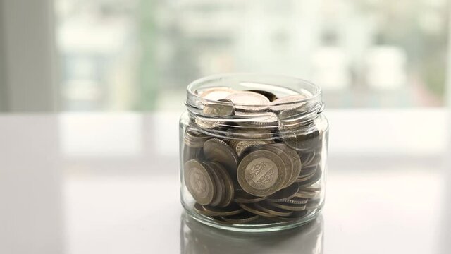 coins falling into the glass jar on wooden table near window, close up. pile of silver coins falls into an empty glass jar on a window background. 