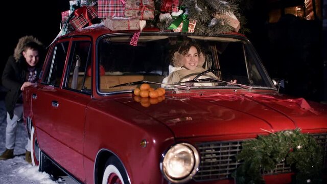 Man pushing Christmas car with woman inside on snowy road at winter night. Woman holding steering wheel stuck car and man pushing car behind