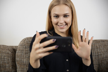Positive young female blogger with phone, wave hand greet friend smiling having videocall online talking on smartphone