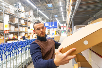 Young man takes a flat box with shelf goods in a large store warehouse.