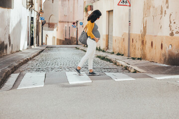 Brunet caucasian pregnant woman walking on the streets of an old city and crossing a crosswalk. Fashionable woman wearing sunglasses, stripes shirt, denim and mustard jacket, looking down. © BASILICOSTUDIO STOCK