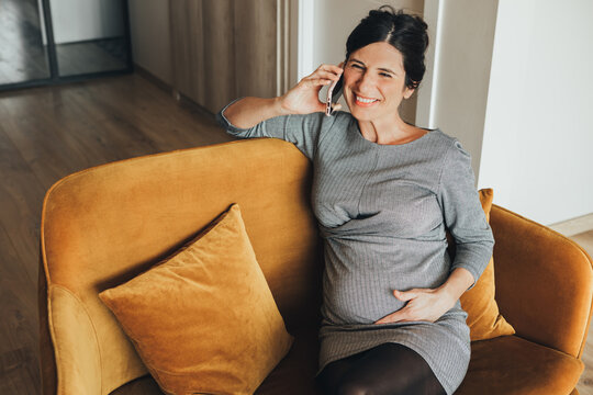 Top View Of Smiling Caucasian Pregnant Woman In Black And White Dress, Sitting In A Mustard Velvet Sofa, Talking On The Phone. Stylish Expecting Mother Having An Interview. Businesswoman Maternity.