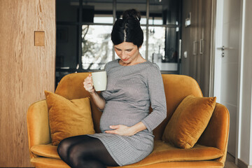 Beautiful caucasian brunet pregnant businesswoman in black and white dress, sitting in a Scandinavian sofa mustard velvet, drinking a cup of tea or coffee and touching her belly. Maternity.