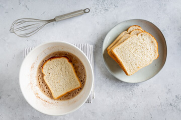 Making process of French toast, one slice of bread soaking in bowl with plate of bread next to it on white grey background, minimal flatlay with copyspace