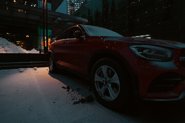 Red car covered in snow parked near modern office building at night. Auto stands in front of modern...