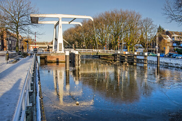 Delft, The Netherlands, February 13, 2021: white steel drawbridge reflecting in the flakes and broken ice on Rijn-Schie canal on a sunny day in winter