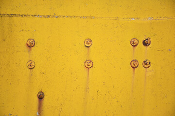 A metal surface painted with yellow paint cracked from age. Rusty steel rivets with rust stains.