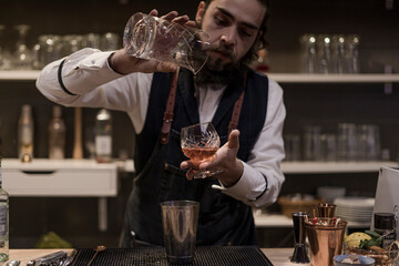 Barman in shirt and apron making an alcoholic drink with ice in a cocktail glass
