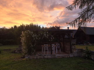 View of summer garden with tiber wooden gazebo pergola with blooming climbing plant flowers and white furniture and surrounding sandstone wall, green grass and bush background and vivid sunset sky.