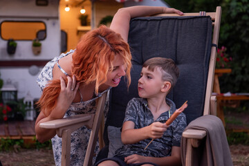 Happy family at picnic near the trailer track. Young mother and her son are sitting on a wooden deck chair, fooling around, eating sausages fried on a barbecue. Vacation outside the city, fun holidays