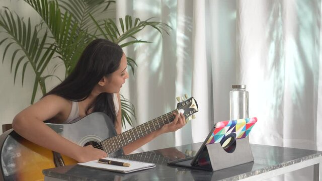 Young Asian woman practicing and learning how to play guitar on laptop tablet.Female guitarist watching online guitar tutorial.Concept of online guitar classes, practicing at home