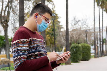 Fototapeta premium Stock photo of young boy wearing face mask due to covid19 using his phone and listening to music in the park.