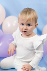 A beautiful little girl 1 year old with blue eyes and white turtleneck sits among the balloons and looks at the camera