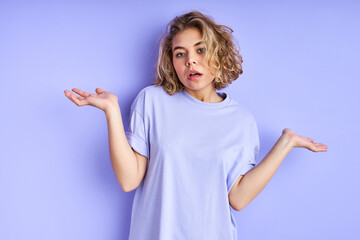 Fototapeta premium unsure female model shrugs shoulders with uncertainty isolated in studio, makes questioning gesture, has clueless and confused look, poses alone against purple wall. Who cares