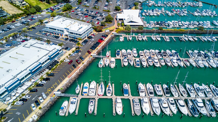 Aerial view on a marina with boat and yachts resting on calm water and cars parked along the shore on a busy plaza. Auckland, New Zealand.