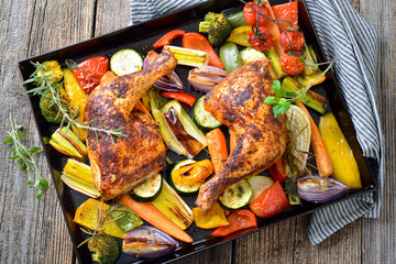 Zwei knusprig gebackene Hühnerkeulen mit mediterranem Gemüse heiß auf dem Backblech serviert –...