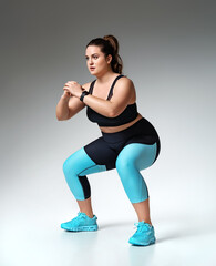 Sporty woman doing squats. Photo of model with curvy figure in fashionable sportswear on grey background. Sports motivation and healthy lifestyle