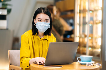 Fototapeta na wymiar Asian female freelancer or student using a laptop for remote work or distance learning sits at the cafeteria with a cup of coffee, wearing medical mask answers emails,browsing internet