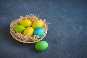 Yellow green and blue easter eggs in wooden plate decorated with decorative shavings on dark grey background. Copy space