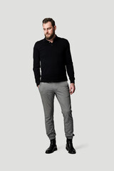 Handsome guy with a beard looking sad with his face away from the camera and a hand in his pocket, wearing black long sleeved shirt and grey pants standing against a grey background.