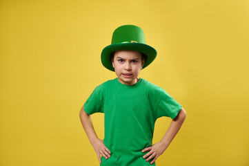 Angry boy wearing leprechaun green hat poses to the camera on a yellow background. Saint Patrick's Day. Copy space