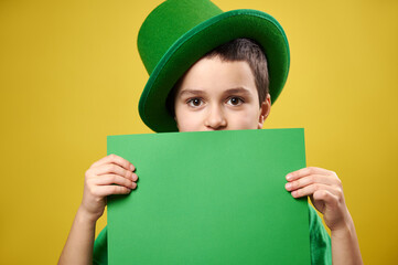 Handsome boy dressed in a leprechaun with a green Irish cap covers his face with a sheet of green...