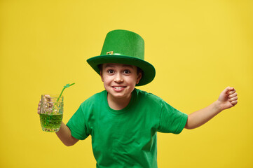 Happy boy in leprechaun hat with hands up holds a glass with green drink and express happiness celebrating the Saint Patrick's Day. Copy space. Yellow background