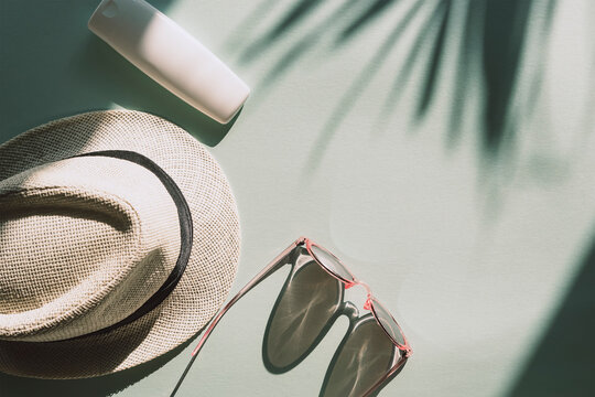 Sun hat, glasses and spf cream or lotion unbranded package on light blue background with palm tree leaf shadow, top view