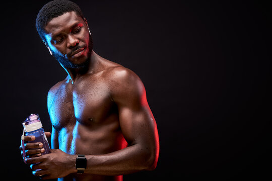 frican shirtless man holding bottle of fresh water after hard intense physical activity, isolated on black background
