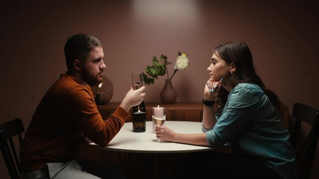 Romantic young couple enjoying talking and sitting together at dinner table in cozy dark living room. Love couple celebrating anniversary or Valentines day having romantic dinner, side view.