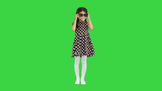 Dark-haired little girl in polka-dot dress putting on black sun glasses on a Green Screen, Chroma Key.