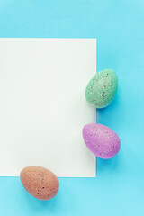 Fototapeta premium Three painted eggs and blank sheet on blue background, top view, flat lay
