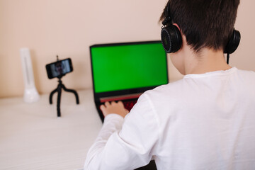 Back view of young boy play gamer on laptop greenscreen. Steamer in earphones capture video on his phone on tripod