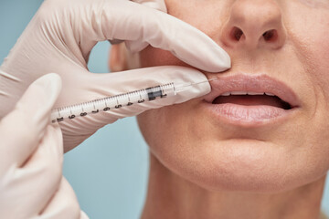 Beauty procedure. Close up of beautician in white gloves making hyaluronic acid injection in lips for adult lady