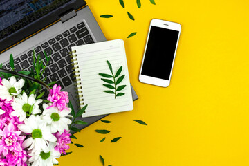 Spring office background, workspace with bouquet of flower, yellow desktop with laptop, blank notepad and smartphone, copy space