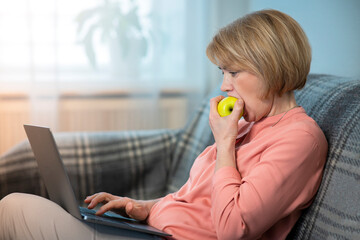 Beautiful woman, elderly senior lady freelancer is working on her laptop computer and eating biting healthy fruit - apple, sitting at home in living room. Distant freelance job, work concept