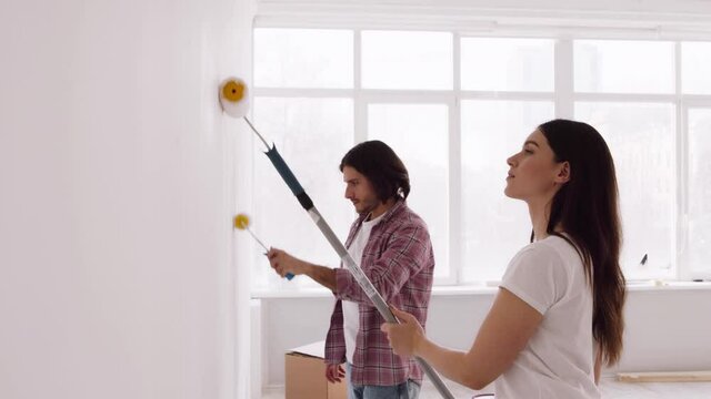 Young married couple making repair together, painting white wall in empty apartment with rollers