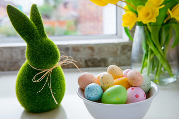 White bowl with colored easter eggs, bouquet of yellow tulips and daffodils flowers and green bunny...