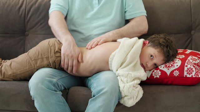 father gives his son a back massage by putting him on his lap. A parent massages the child's back while sitting on the couch at home.