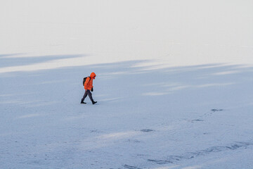 A man in an orange jacket walks in the snow on a frozen lake.