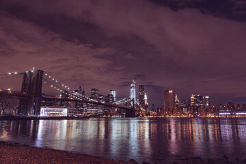 Fototapeta na wymiar Illuminated Brooklyn Bridge And Buildings By River Against Sky At Night in New York City