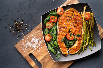 grilled salmon steak, asparagus and fresh spinach with tomatoes on a black square plate on a dark background