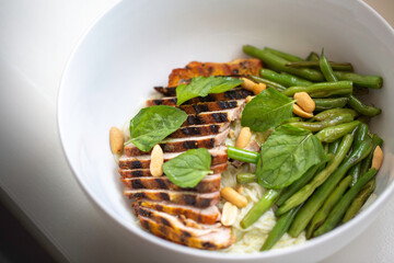 Sliced grilled chicken and string bean with peanuts and mint leave on a noodle in a white bowl. Diagonal top view.