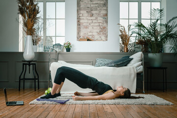 A young woman goes in for sports at home, online workout . The athlete  pumps up the buttocks, makes the gluteal bridge in the bedroom, in the background there is a bed, a vase, a carpet.