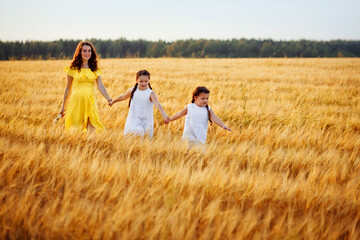 A pregnant woman in a yellow dress with two daughters in white sundresses walks in a wheat field, holding hands, at sunset