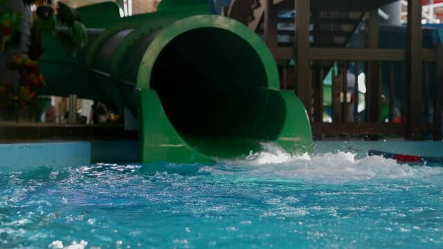 A little girl slips out of the pipe of a green water slide in a water park, she plops into the pool on an inflatable circle.