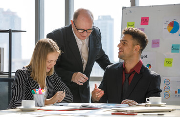 Adults caucasian business 2 men and 1 woman are meeting about the plan and have a white coffee cup, notebook put on table and whiteboard are backward of them with more paper note on it in the morning