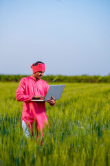 Young indian farmer using laptop at green wheat field