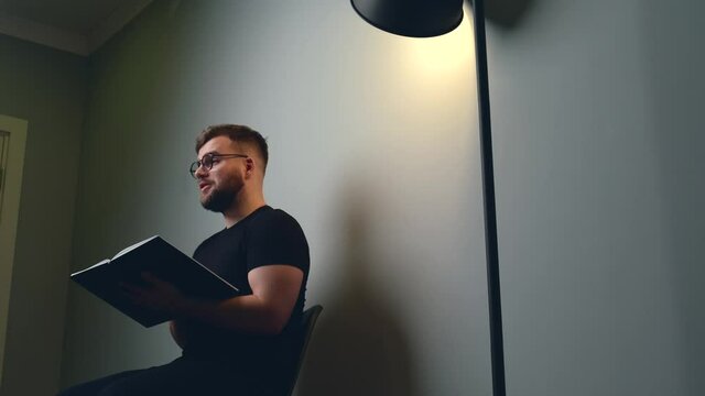 Caucasian man with beard is having online courses on a gray studio wall speaking to camera from a book